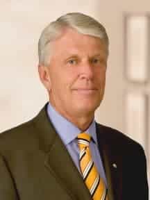 A man with short gray hair is wearing a dark suit, a light blue shirt, and a yellow and black striped tie. He is standing indoors with a neutral expression and a blurred background.