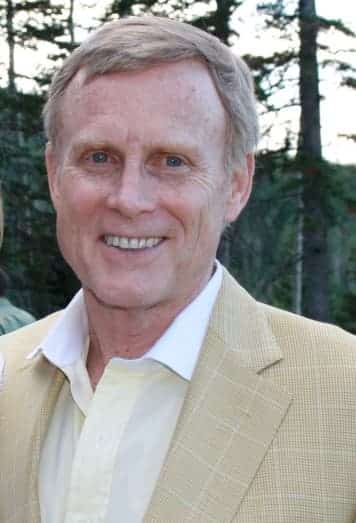 A person smiling outdoors, wearing a light yellow shirt and a beige jacket. There are trees in the background, suggesting a natural setting.