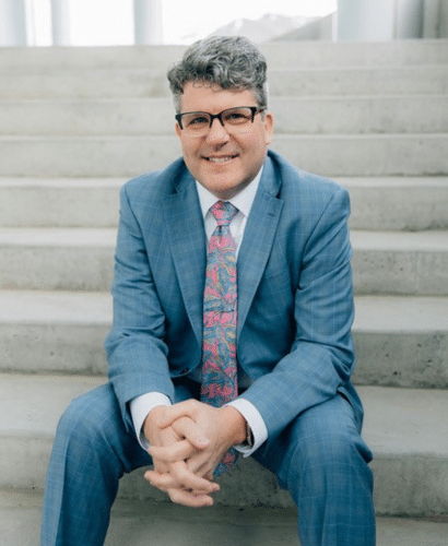 A man in a blue suit with a colorful patterned tie sits on outdoor concrete steps, smiling and clasping his hands in front of him. He has short, curly gray hair and glasses.