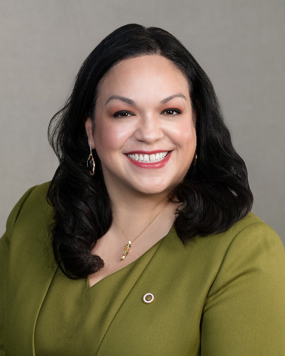 A woman with long dark hair smiles warmly, wearing a green blazer, gold jewelry, and drop earrings, posed in front of a plain light gray background.