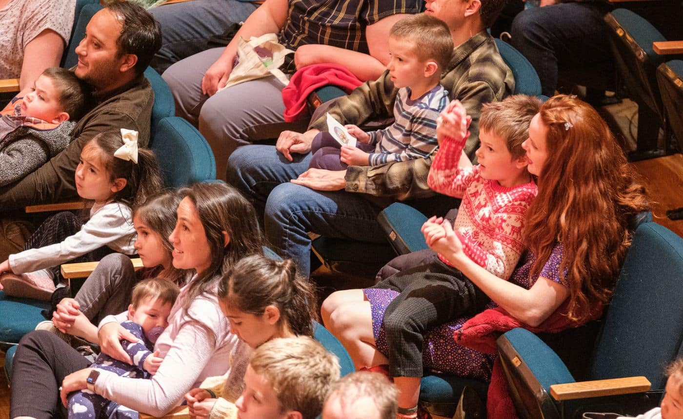 A group of adults and children sit closely together in theater seats, watching a performance. The children appear excited and engaged, some sitting on adults laps. The audience is dressed casually.