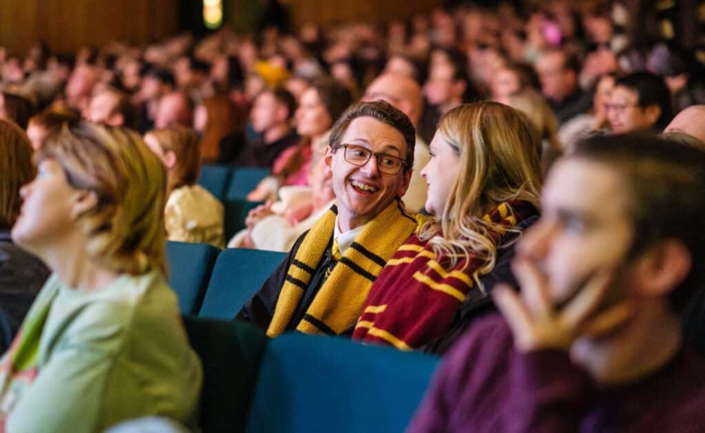 A man wearing glasses and a yellow scarf smiles and talks to a woman in a red and yellow scarf. They are seated among a large audience in a theater or auditorium.