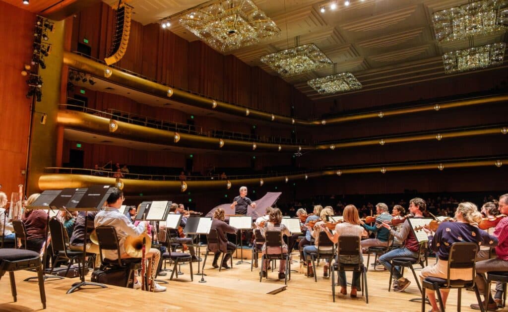A symphony orchestra rehearses on stage in a large, elegant concert hall with three balconies, golden accents, and sparkling chandeliers overhead. The conductor stands in front of the musicians, who are seated facing the stage.