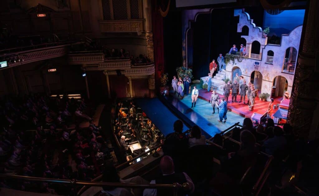 A wide view of a theater stage shows actors performing in front of a set resembling Mediterranean architecture, as an orchestra plays below and an audience watches from balconies and seats.