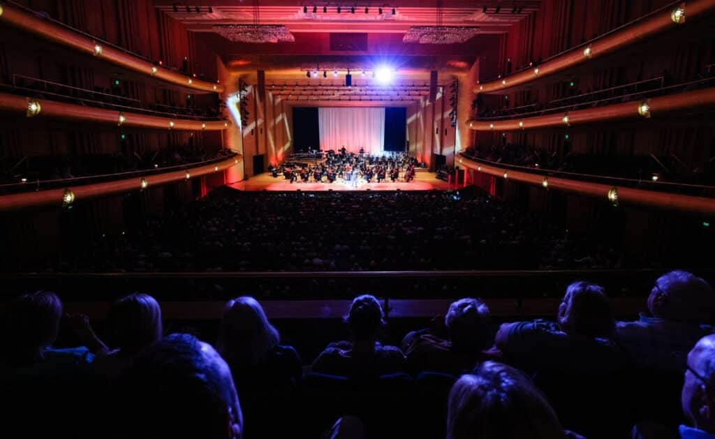 A large audience watches an orchestra performing on a brightly lit stage inside a grand concert hall with balconies and high ceilings. The room is dim except for the stage lights.