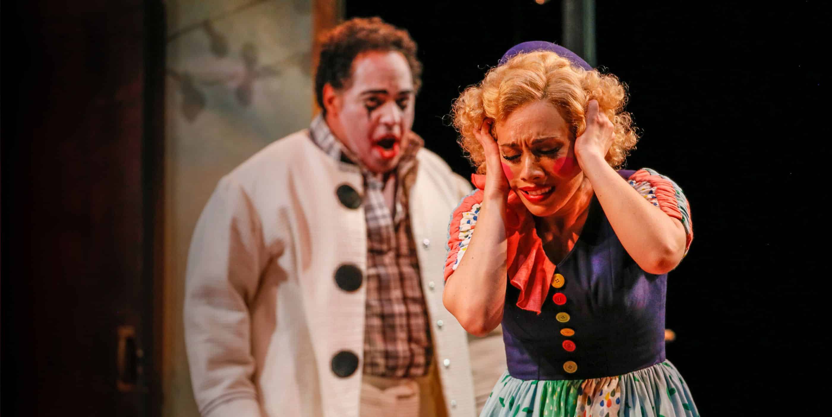 A scene from an opera features a man in a clown costume standing behind a visibly distressed woman in colorful, vintage-style clothing. The woman holds her head with both hands, while the man, with face paint, has a concerned expression.