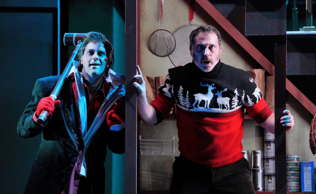Two men act in a theatrical scene. The man on the left holds a rifle and wears a dark outfit with red gloves. The man on the right looks surprised, wearing a festive sweater with reindeer. They are in a room with shelves and cans.