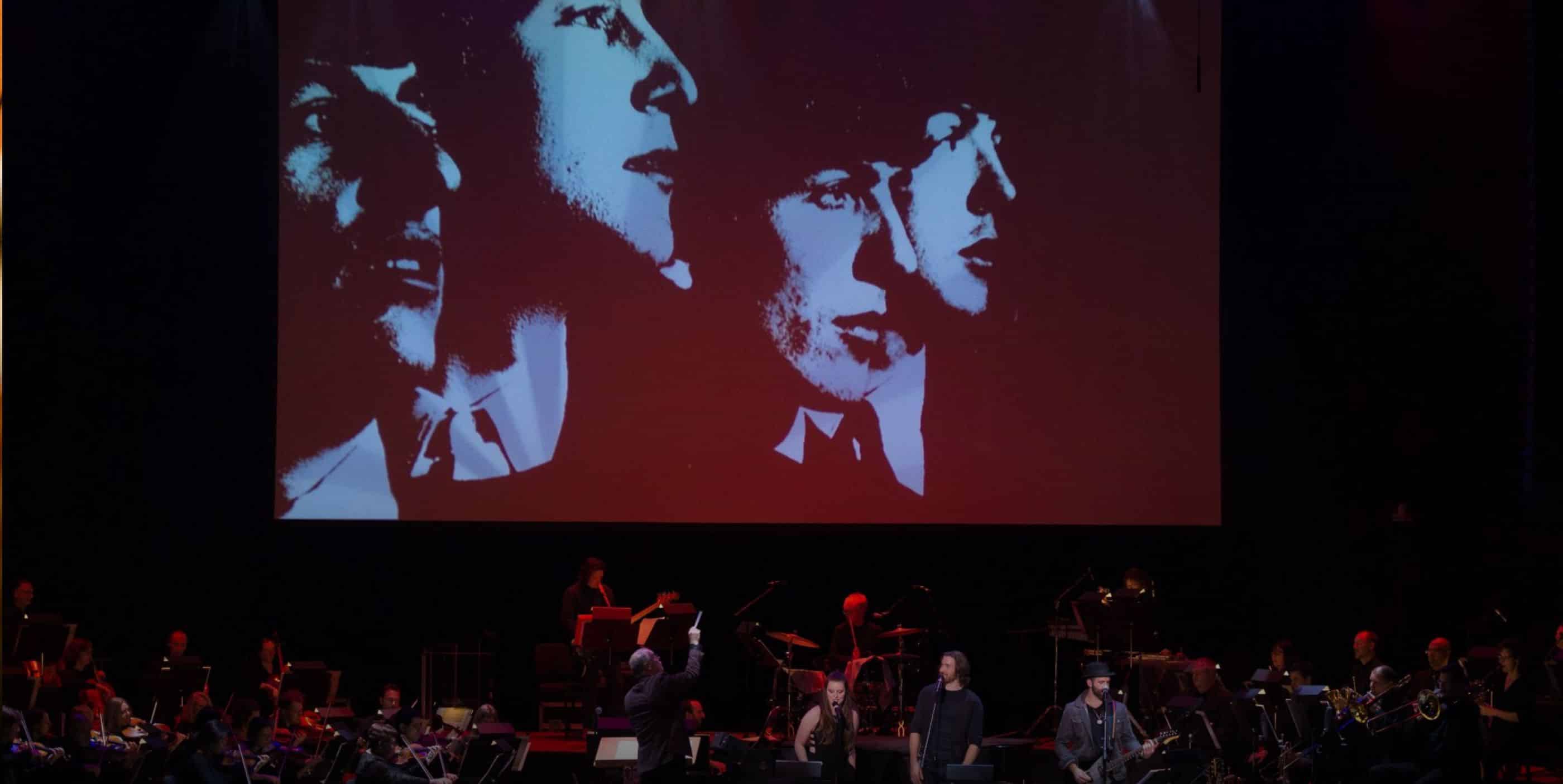 A live orchestra performs on stage. Above them is a large screen displaying a black and red image of four faces. The conductor and musicians are visible, with various instruments in the scene.