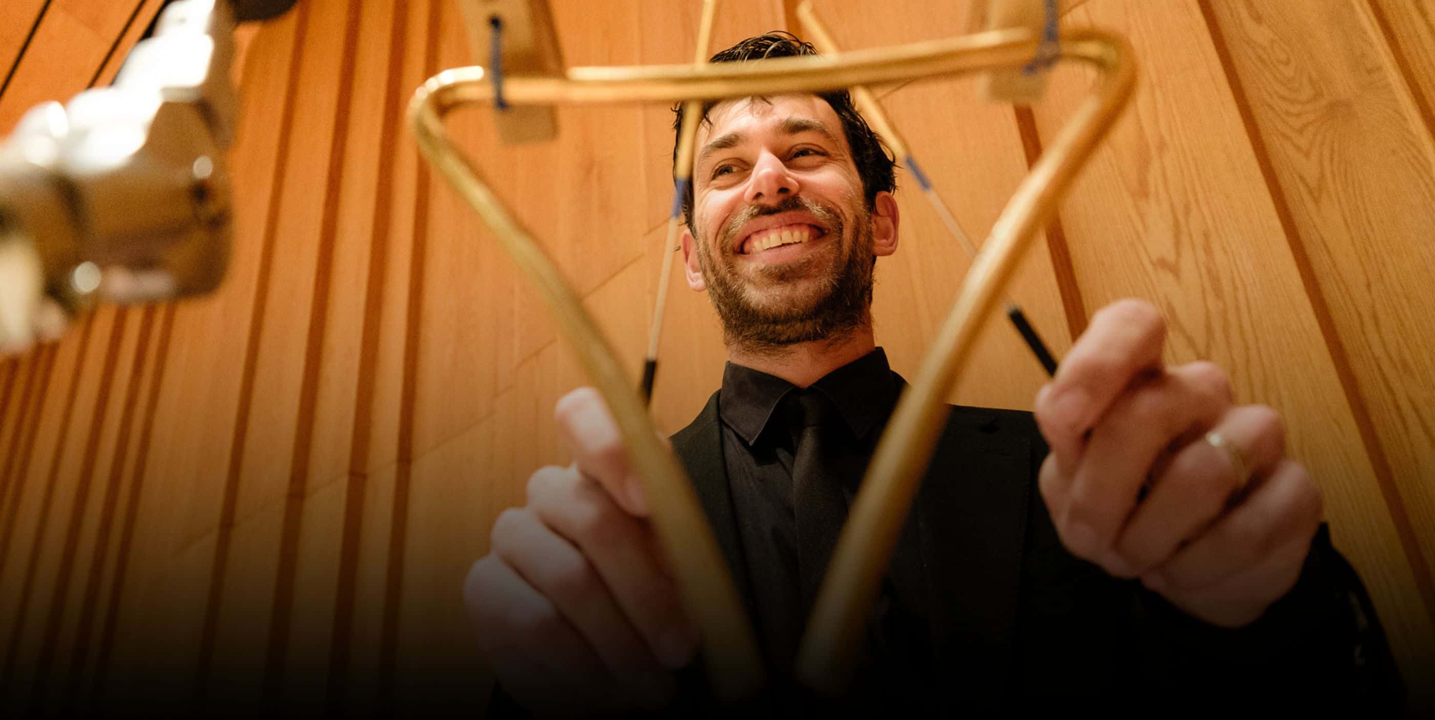 Smiling person playing a triangle instrument in a wooden-paneled room.