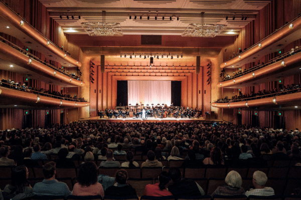 A large audience sits in a grand concert hall with balconies, watching an orchestra perform on a brightly lit stage with a white backdrop. The atmosphere is elegant and the hall is filled with people.