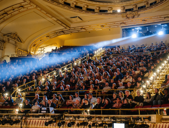A large audience sits in tiered rows inside an ornate theater, bathed in warm lights with a beam of spotlight crossing the scene from left to right. The balconies and decorative ceiling are visible.