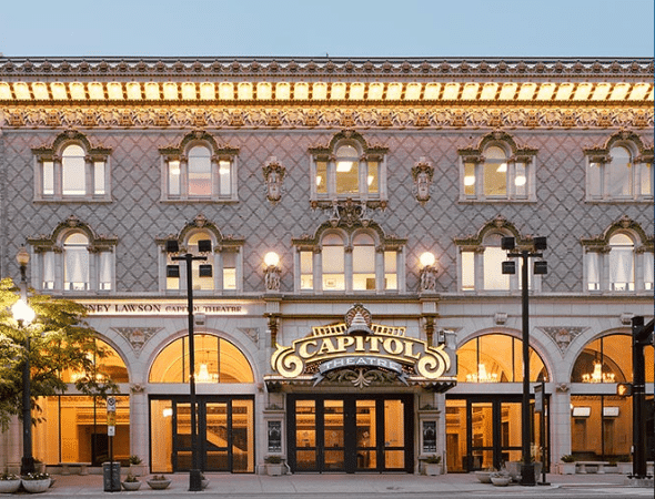 The ornate facade of the Capitol Theatre, featuring arched windows, intricate architectural details, and a large illuminated marquee sign above the entrance, lit by warm lights in the early evening.