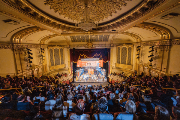 An audience watches a live stage performance in an ornate, historic theater with a grand chandelier, balcony seating, and intricate gold detailing on the walls and ceiling.