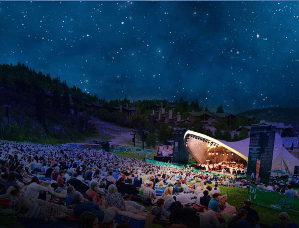 A large crowd sits on a grassy hillside at night, watching an outdoor concert under a starry sky, with musicians performing on a brightly lit stage surrounded by trees and distant buildings.