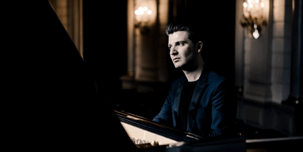 A man in a dark suit plays a grand piano in an elegant, dimly lit room with ornate wall sconces and soft, warm lighting in the background.