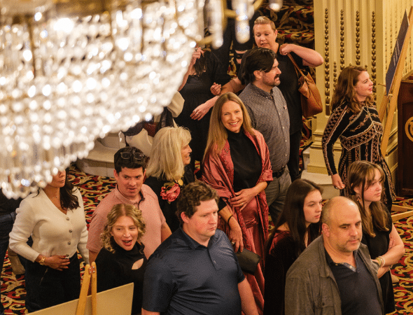 A group of people, dressed in semi-formal attire, stand and walk in a carpeted, ornate room with chandeliers and columns, appearing to attend an event or gathering.