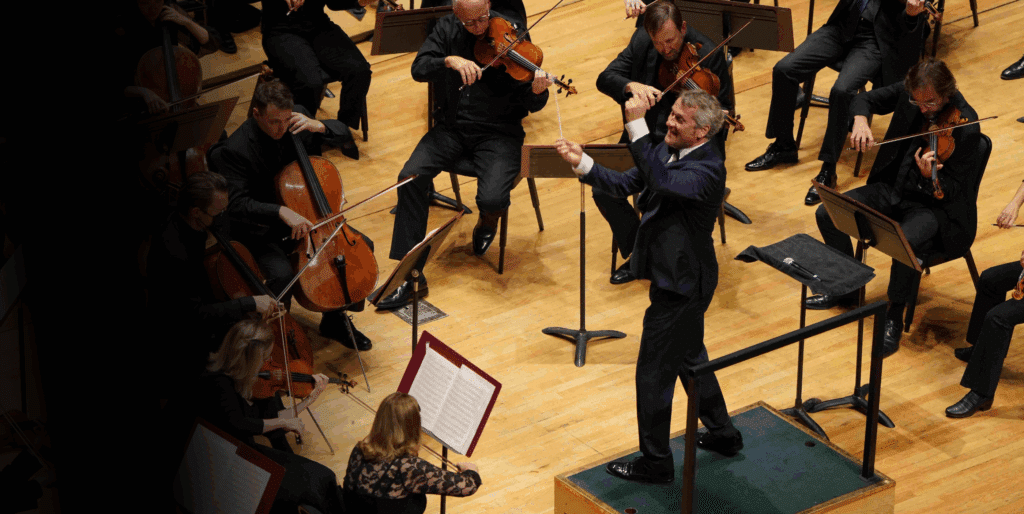 A conductor energetically leads an orchestra of musicians playing string instruments on a wooden stage, with music stands and sheet music visible.