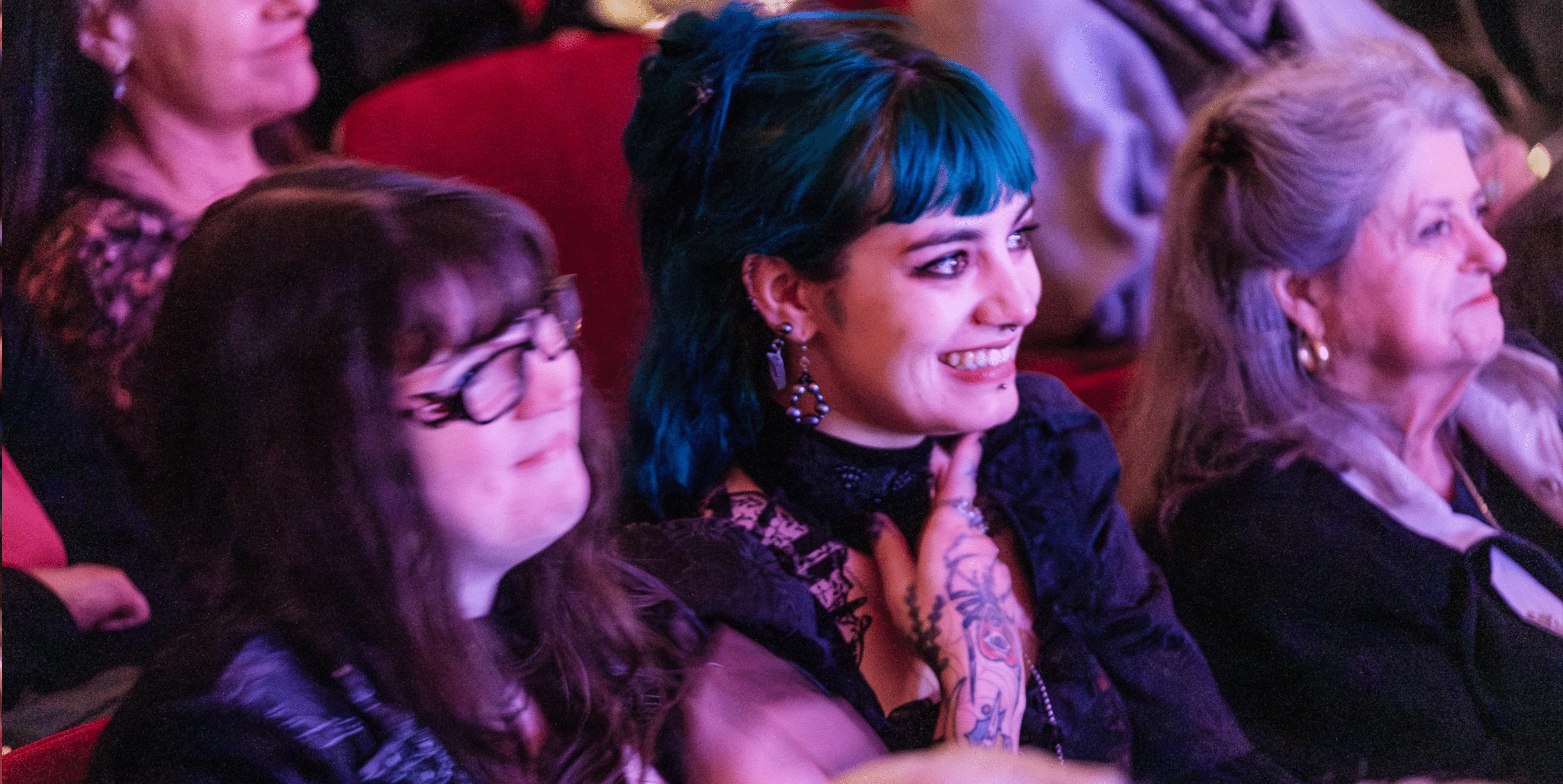 Two women sit closely together in an audience. One has teal hair, visible tattoos, and earrings, smiling brightly. The other has long brown hair and glasses. Both appear engaged, with more people seated around them.