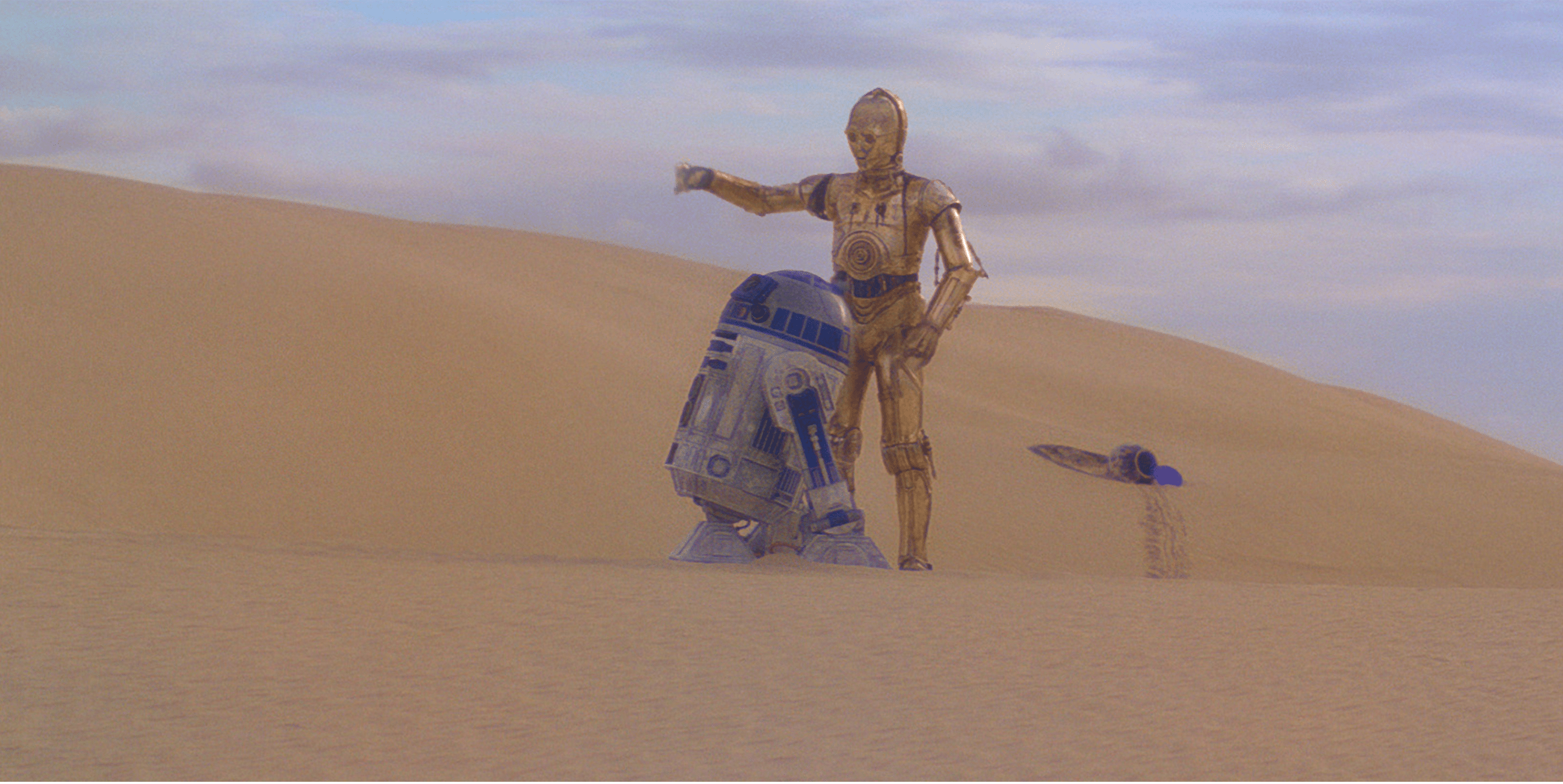 Two robots, a gold humanoid and a blue-and-white dome-topped droid, stand in a vast sandy desert under a cloudy sky. A small escape pod lies partially buried in the sand behind them.