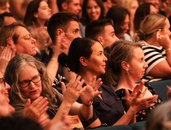 A diverse group of people sitting close together in an audience, smiling and enthusiastically clapping their hands, suggesting they are enjoying a performance or event.