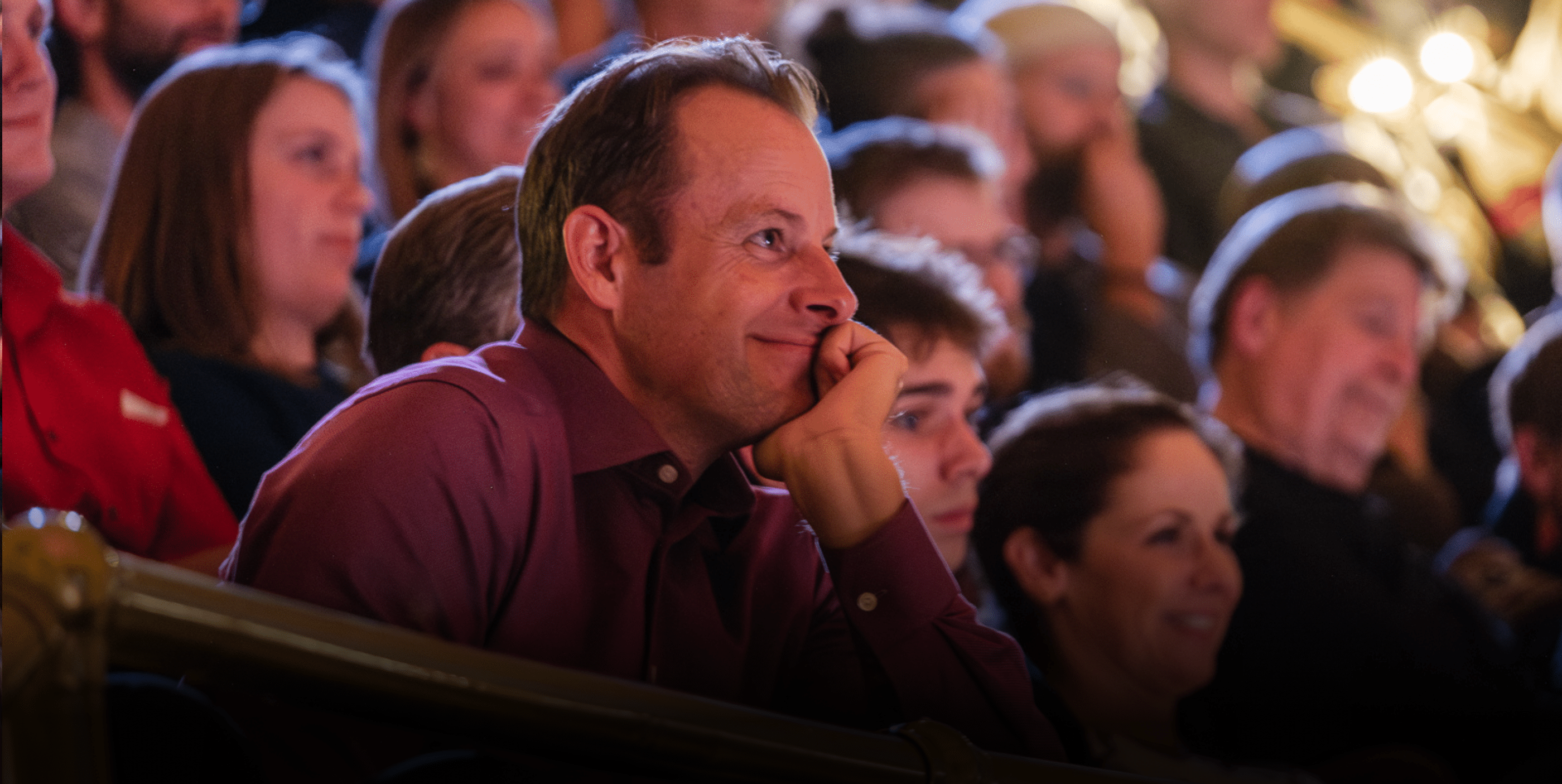 A man in a maroon shirt sits in a crowded theater audience, resting his chin on his hand and smiling while watching the performance. The people around him also appear focused on the stage.