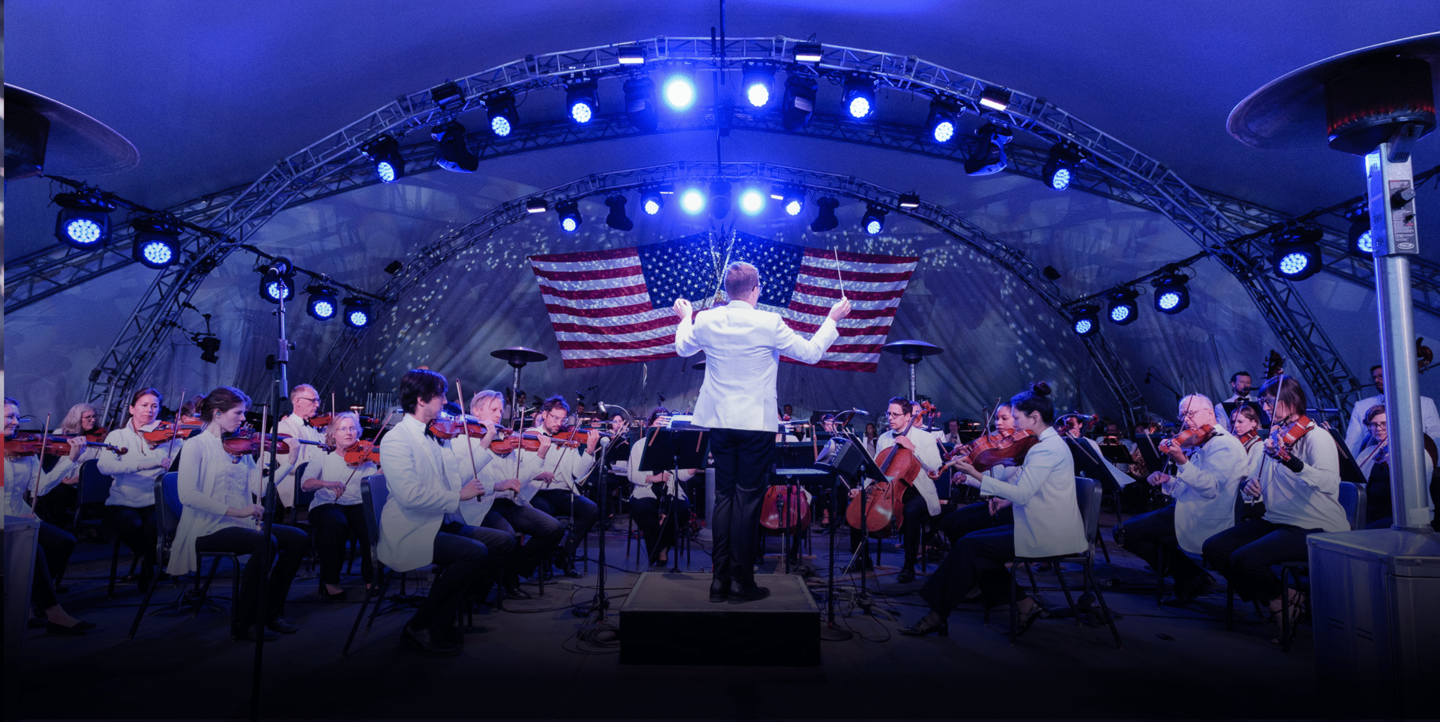 A conductor leads an orchestra on stage under blue lights, with musicians in white jackets playing instruments. An American flag hangs in the background inside a large, curved outdoor structure.