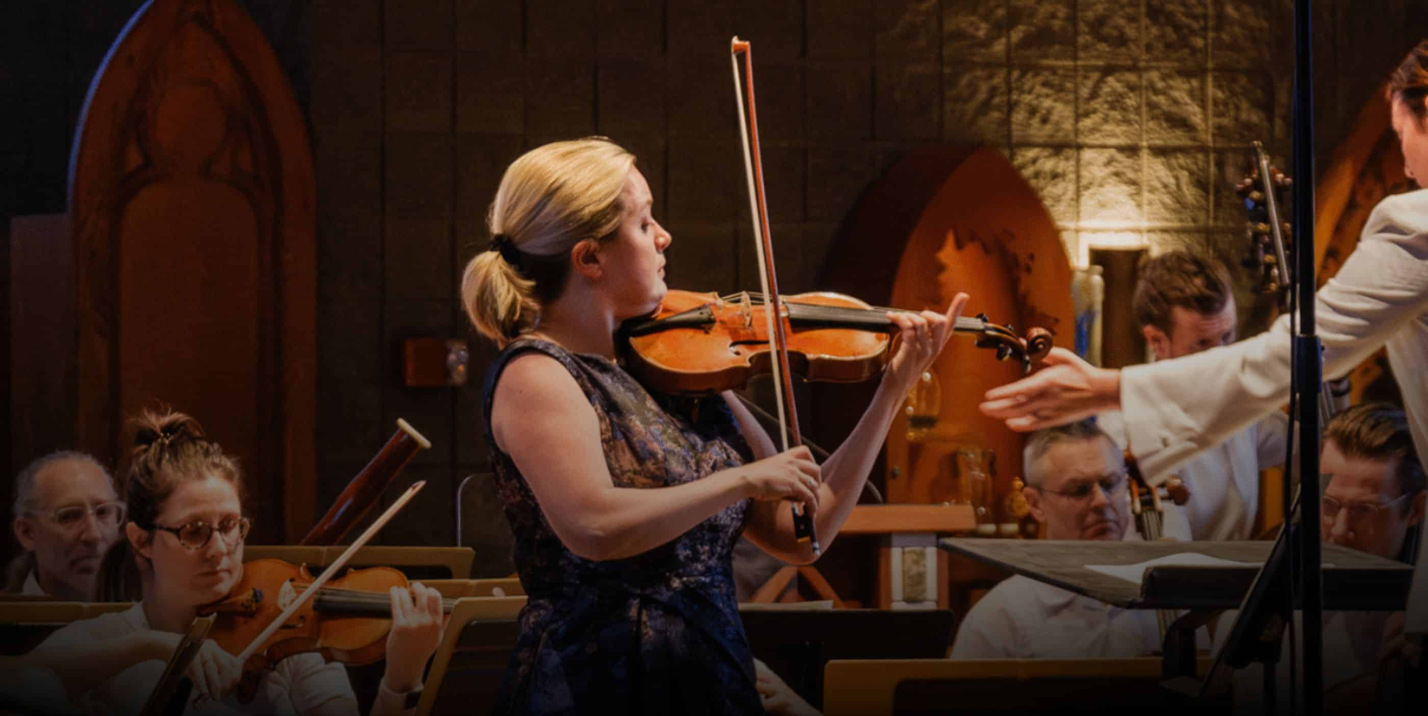 A woman in a dark dress plays the violin on stage with an orchestra, while a conductor extends his hand. Other musicians are seated in the background under warm lighting.