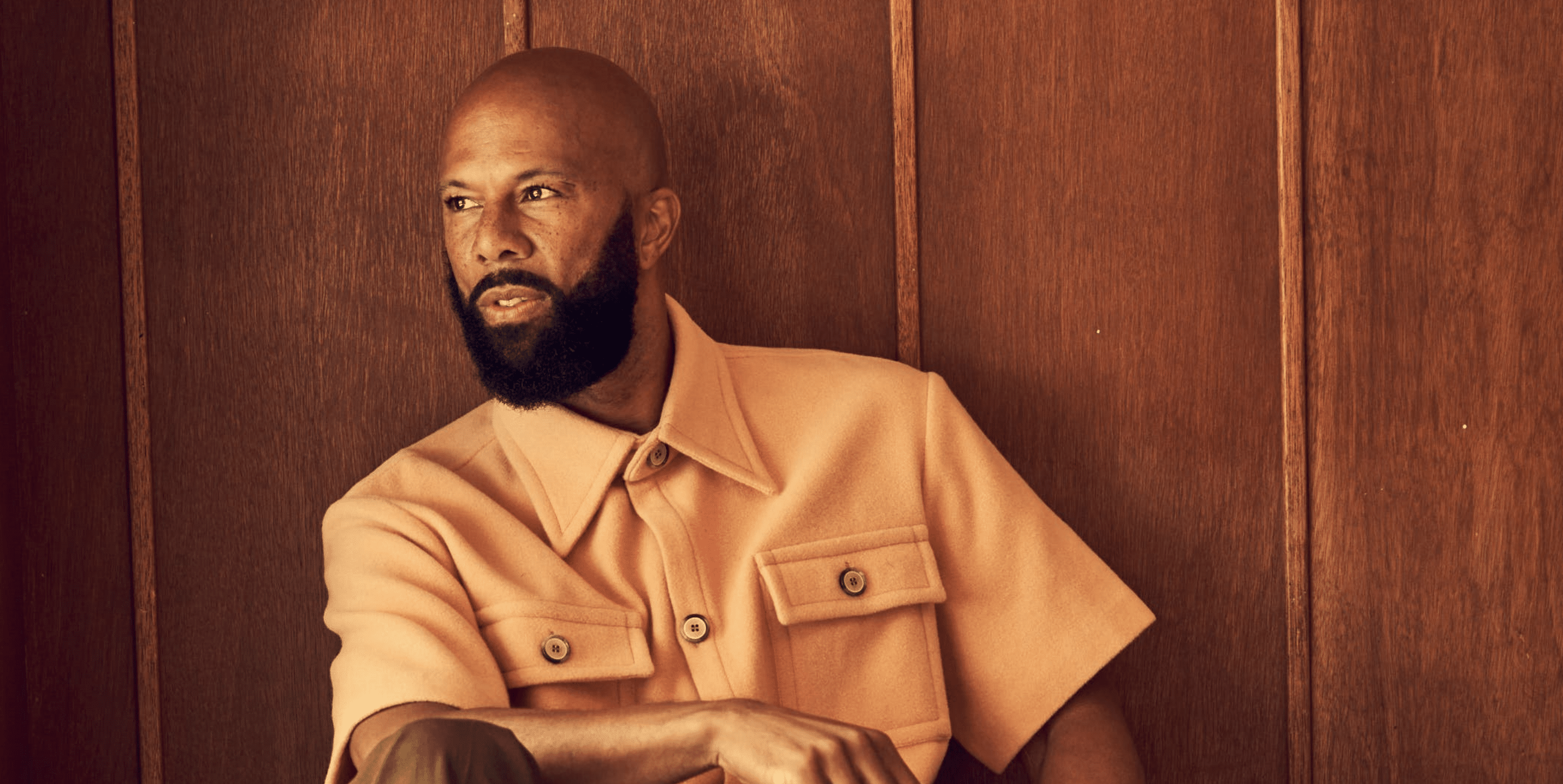 A man with a bald head and full beard, wearing a short-sleeved beige button-up shirt, sits in front of a wooden paneled wall, looking to the side with a thoughtful expression.