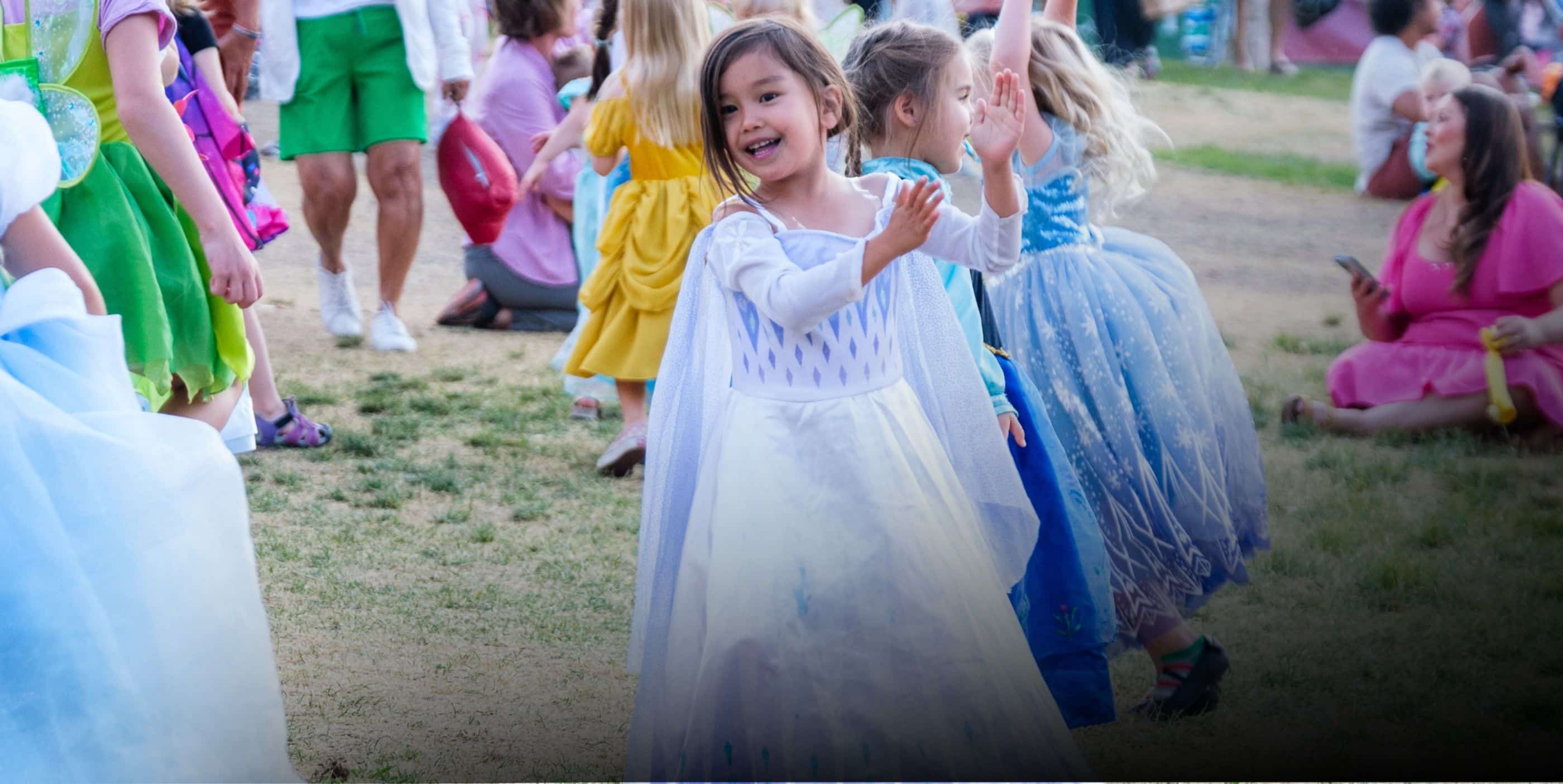 A young girl in a white princess dress smiles and claps her hands while dancing outdoors with other children, some also dressed in colorful costumes, at a playful gathering or celebration.