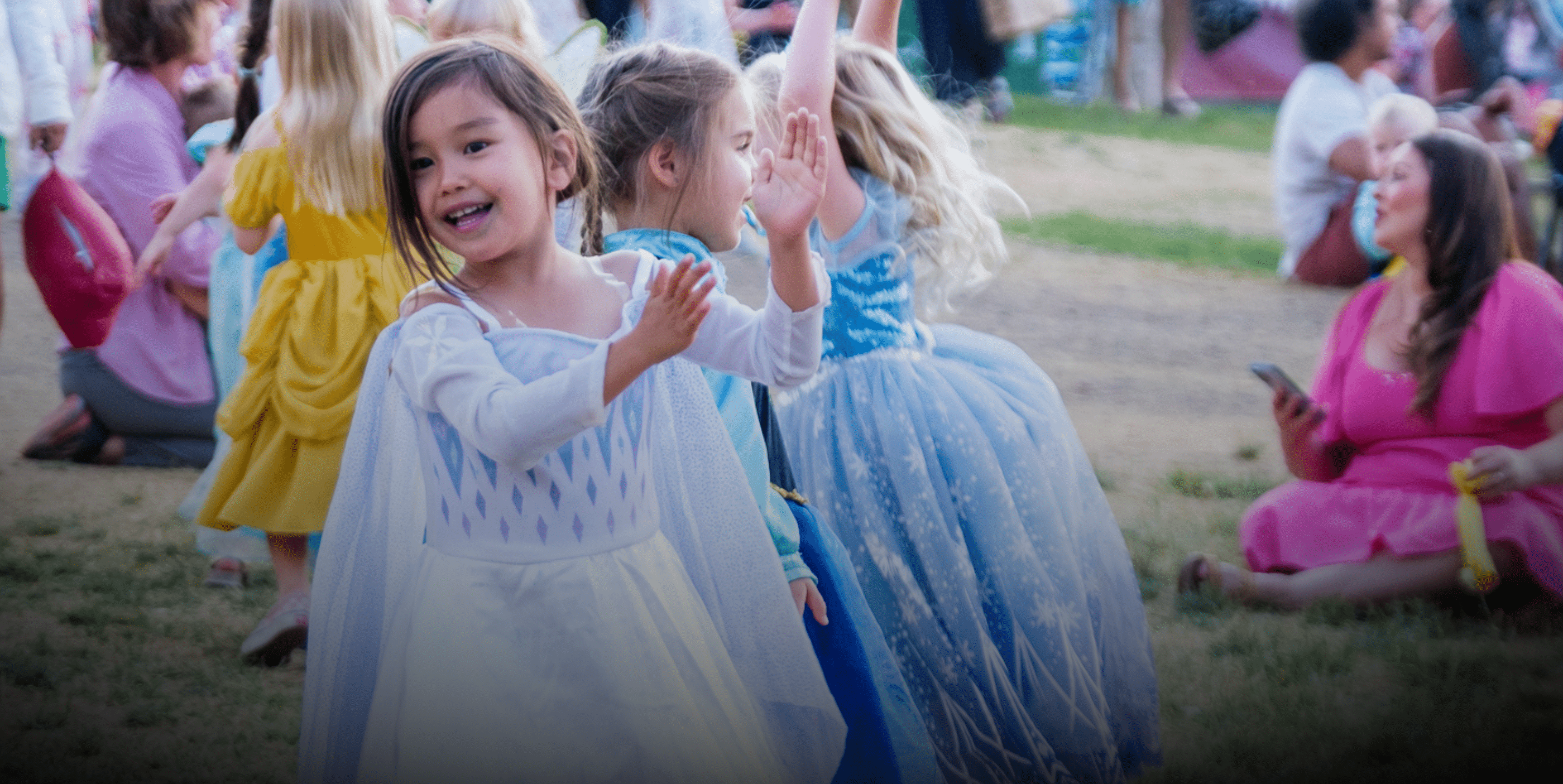 Young children in colorful princess costumes play and dance outdoors at a park event, while adults and other kids sit or stand in the background. One child in a white dress smiles and claps in the foreground.