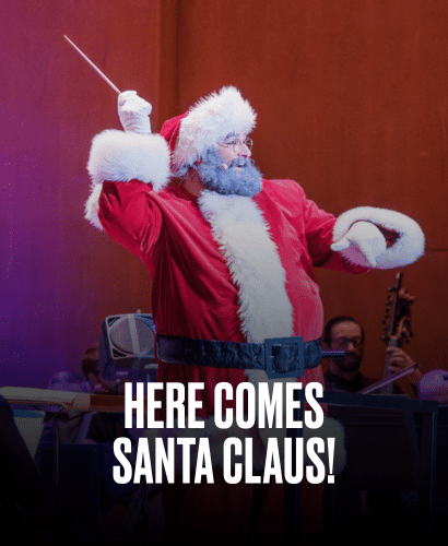 A person dressed as Santa Claus conducts an orchestra onstage, holding a baton with one arm raised. Bold white text at the bottom reads, HERE COMES SANTA CLAUS!.