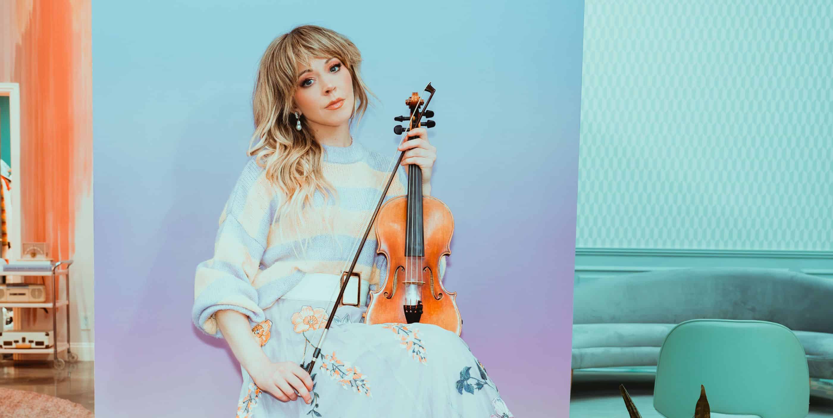 A woman with long blonde hair sits holding a violin. She wears a pastel striped sweater and a floral skirt, with a soft blue and purple background and light-colored modern furniture around her.