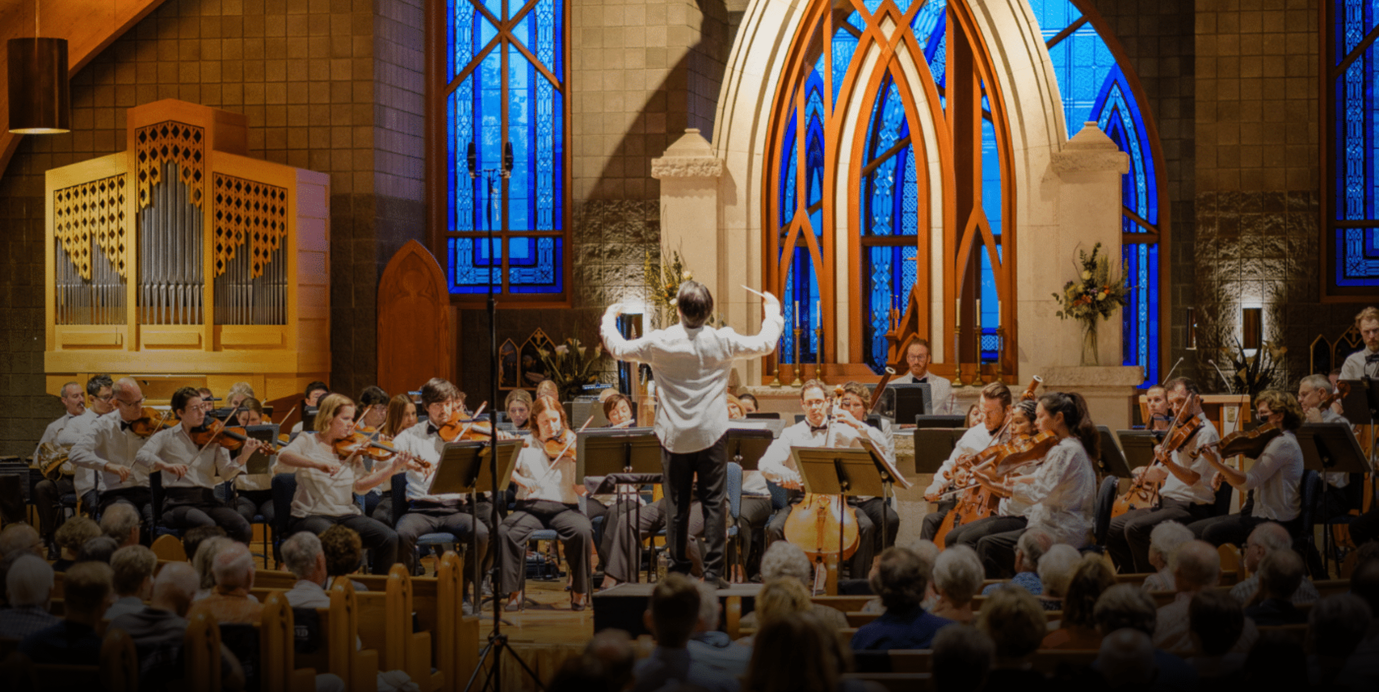 A conductor leads an orchestra performing in a church with blue stained-glass windows, wooden arches, and an audience seated in pews. Musicians play string and wind instruments under warm lighting.