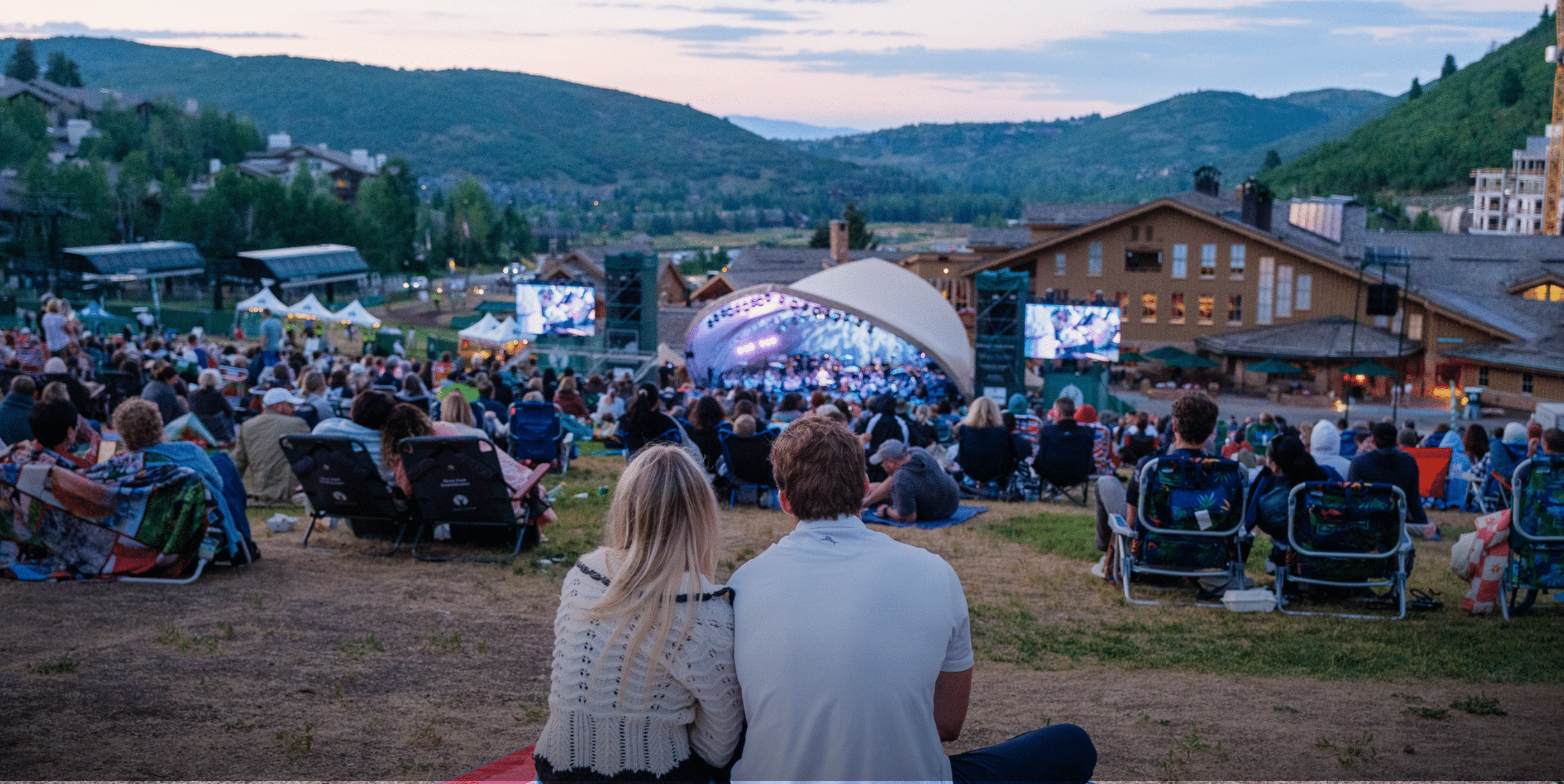 A couple sits on a grassy hill among a crowd at an outdoor concert, watching a band perform on a lit-up stage, with mountains and buildings in the background at sunset.
