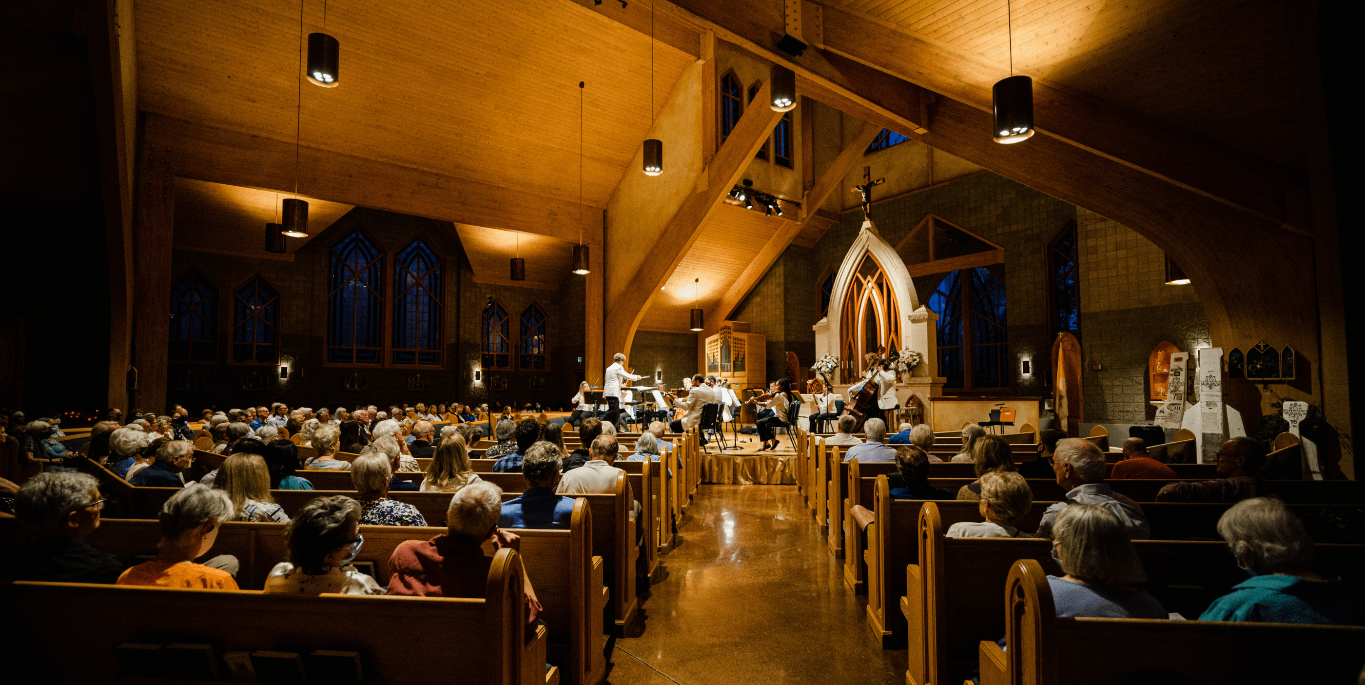 A large audience sits in wooden pews inside a church with high vaulted ceilings, watching a musical performance on a well-lit stage near the altar. The space is warmly lit with stained-glass windows visible.