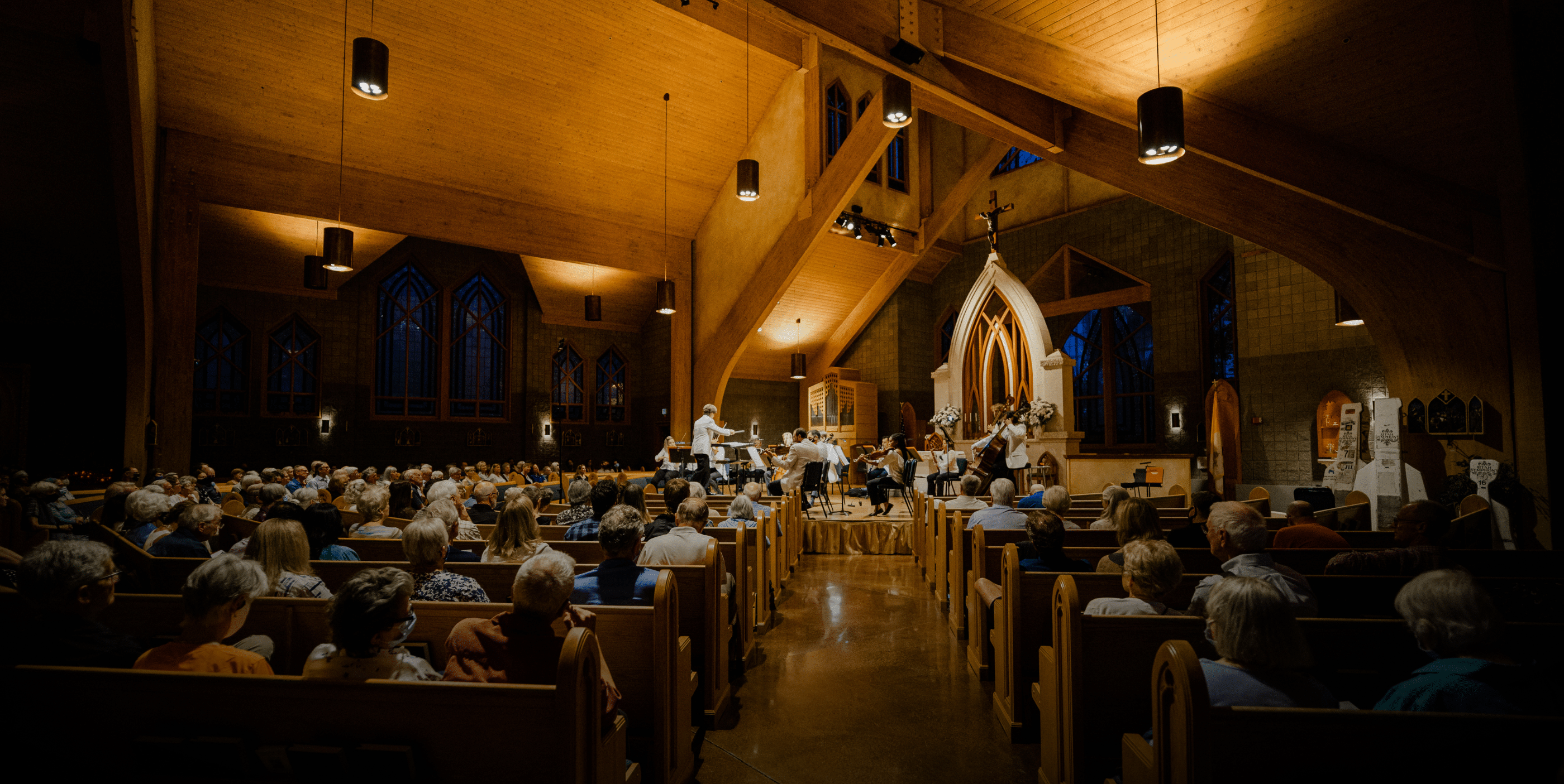 A large audience sits in wooden pews inside a church with high vaulted ceilings, watching musicians perform at the front under warm lighting. Stained glass windows and wooden beams are visible in the background.