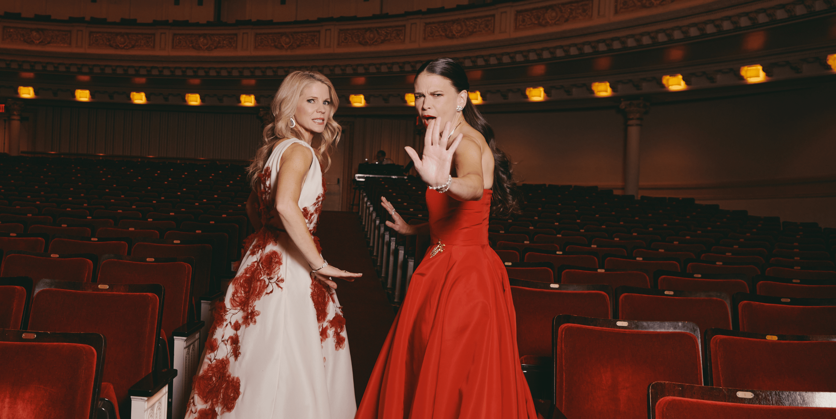 Two women in elegant red and white gowns stand among red theater seats. Both face the camera with playful expressions, one gesturing with her hand as if to stop or direct attention. The theater appears empty.