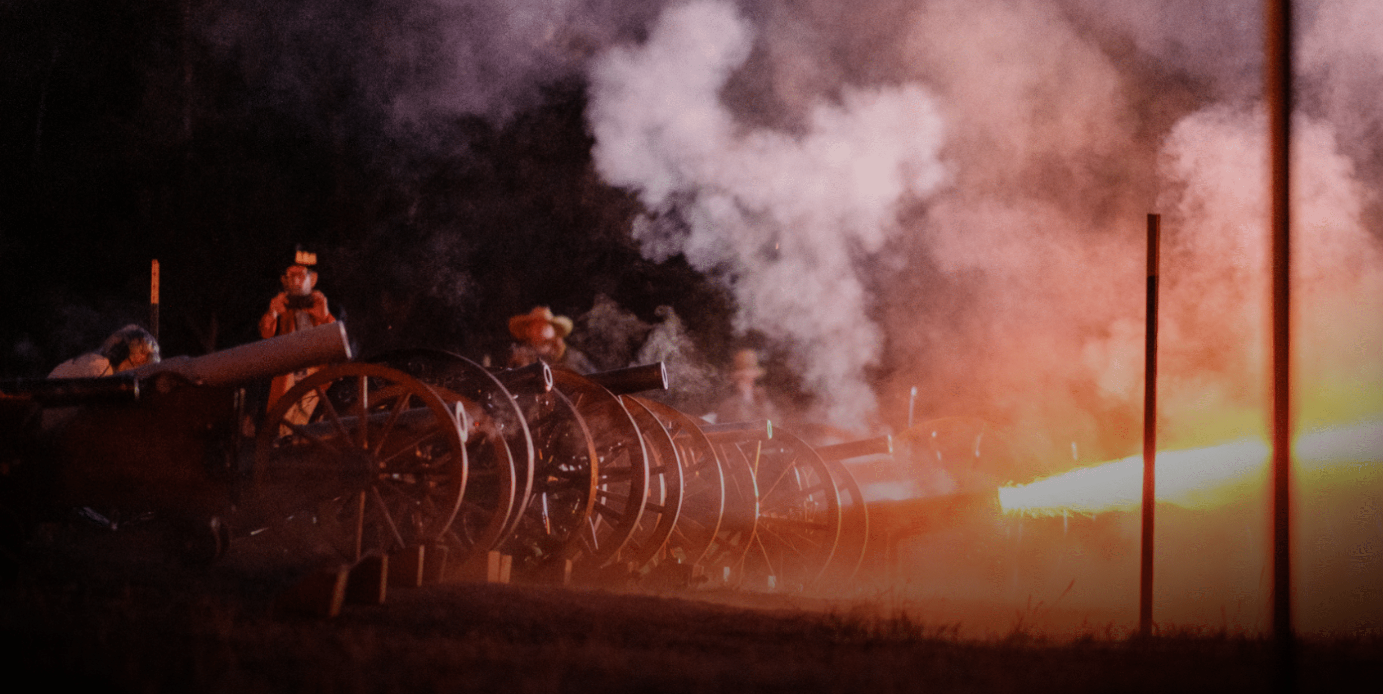 A row of historical cannons is fired at night, producing bright flashes and thick smoke, while people in period costumes stand nearby, illuminated by the light.
