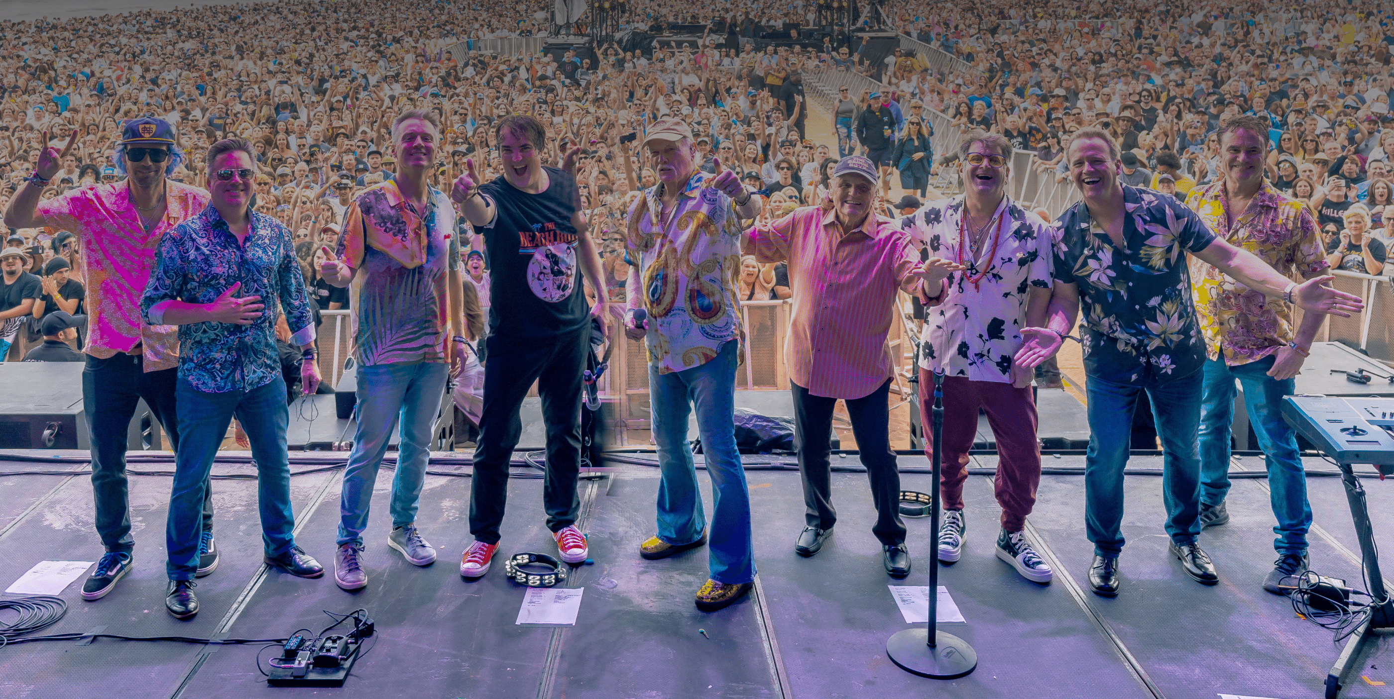 A band of nine smiling men stands on stage in colorful outfits, posing for a photo with a massive cheering crowd behind them at an outdoor concert. Microphones and equipment are visible on the stage.