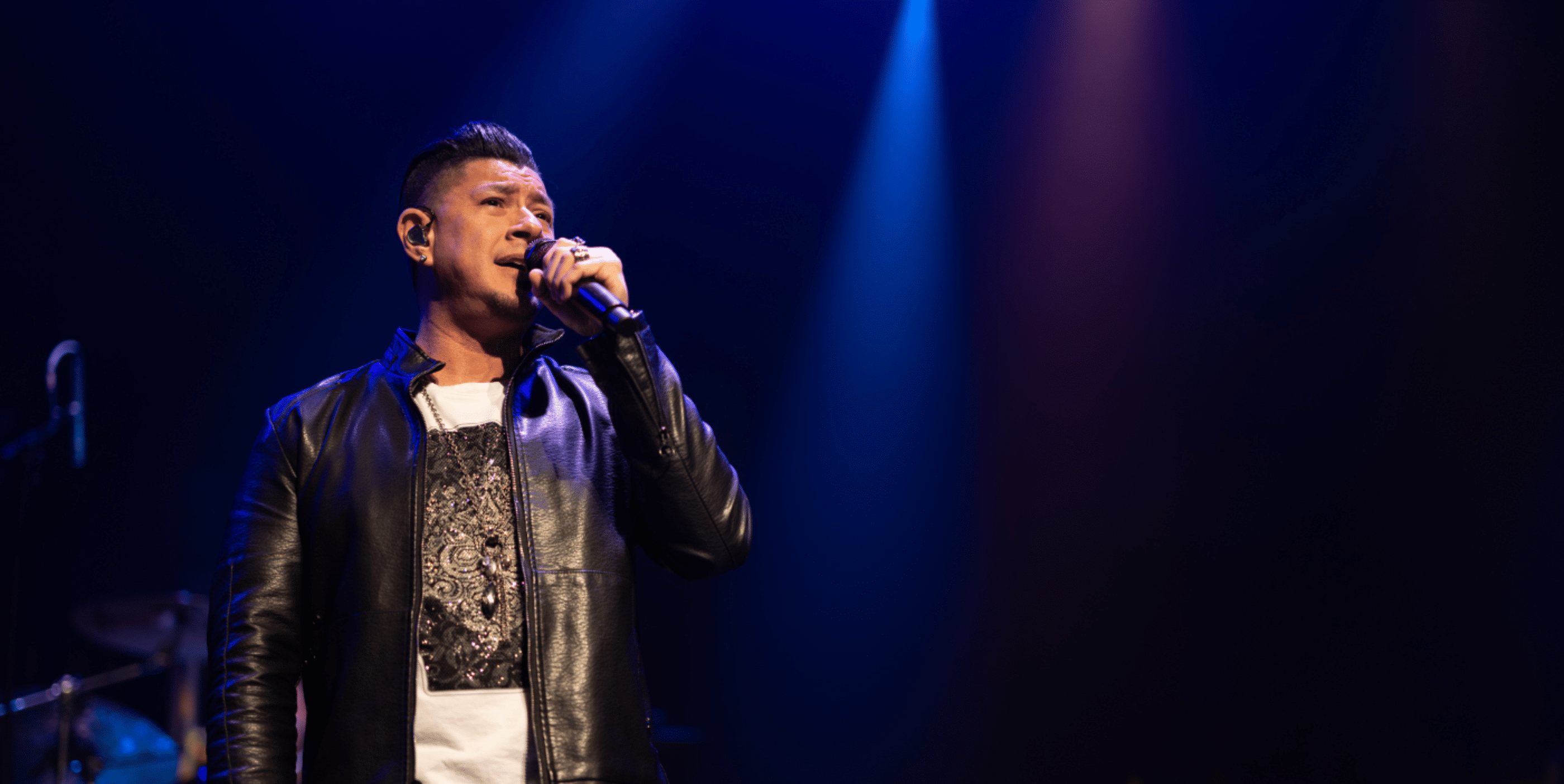 A man wearing a black leather jacket sings into a microphone on stage under blue and purple spotlights, with a dark background and musical equipment faintly visible.
