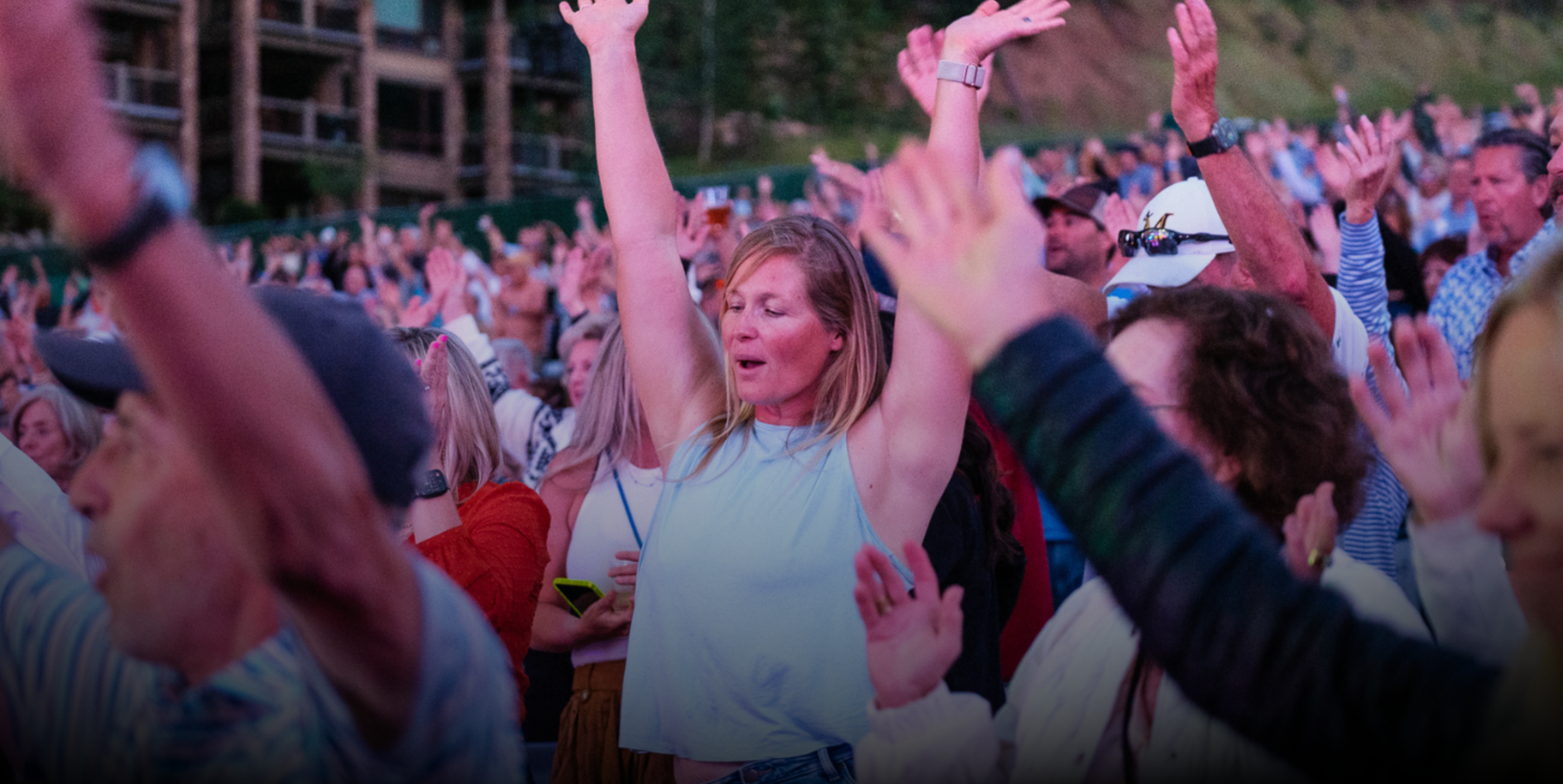 A crowd of people with their arms raised, enjoying an outdoor event. A woman in the center stands with eyes closed and hands up, surrounded by others also celebrating. Trees and buildings are visible in the background.