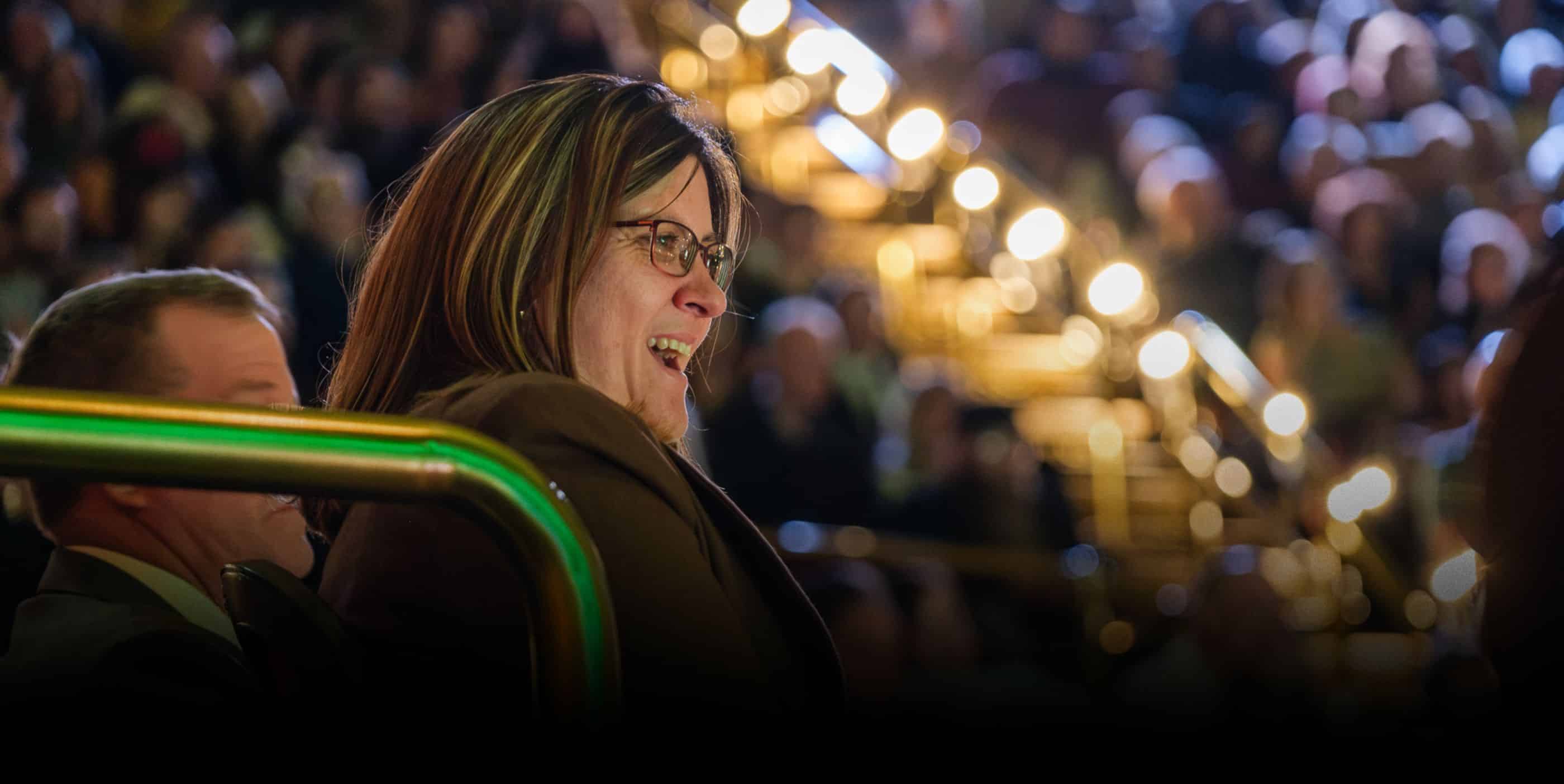 A woman wearing glasses smiles while seated in a crowded, brightly lit auditorium with blurred lights and people in the background.