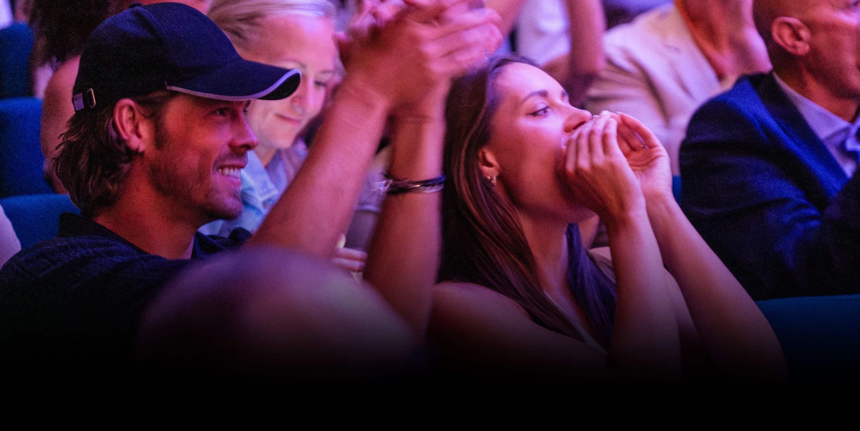 A man in a baseball cap and a woman are cheering and clapping enthusiastically in a crowded audience. The scene is brightly lit, and other people are also visible in the background, joining in the applause.