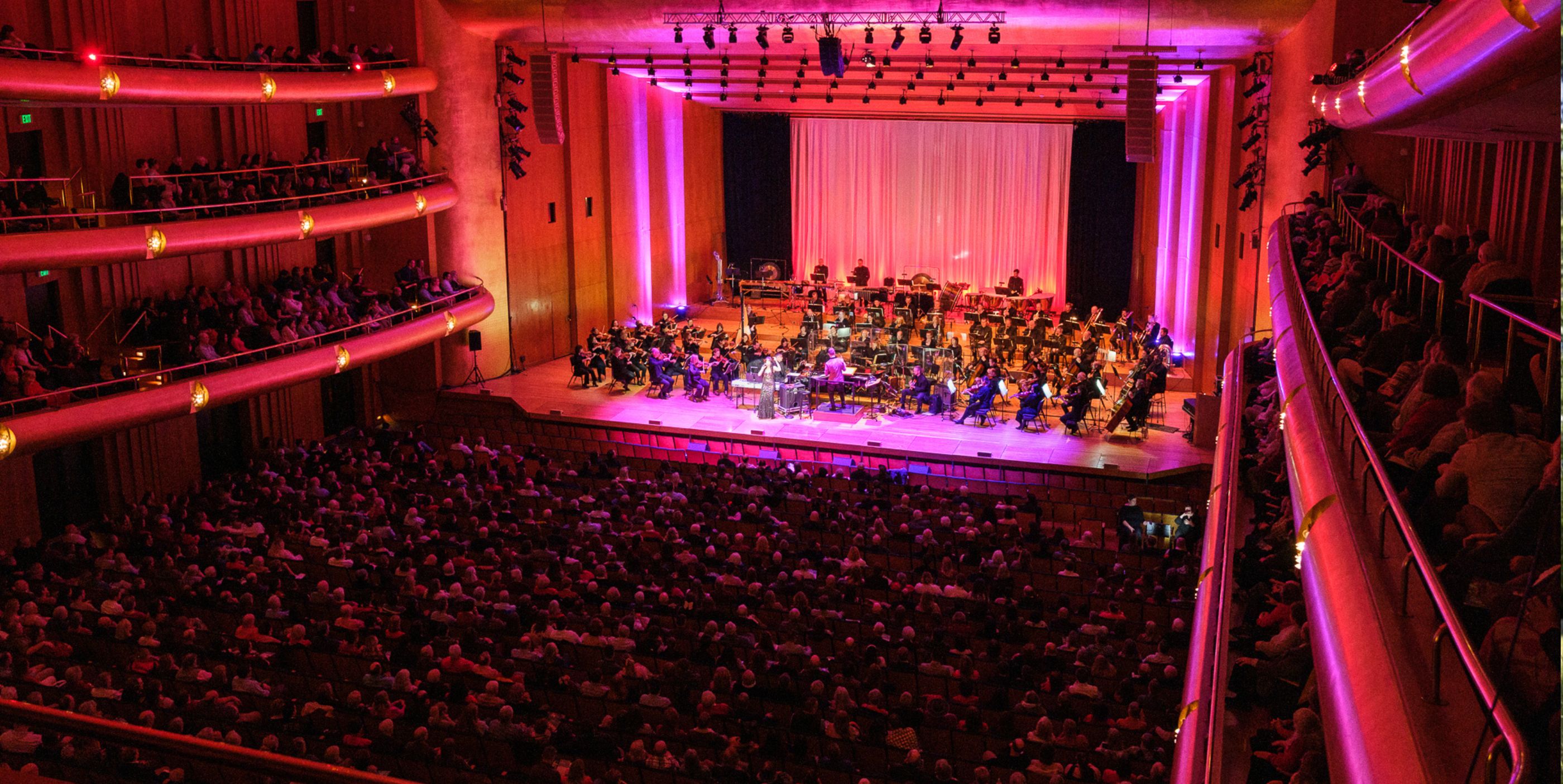 A large audience watches an orchestra perform on stage in a modern concert hall with pink lighting, tiered balconies, and a full house of seated attendees.