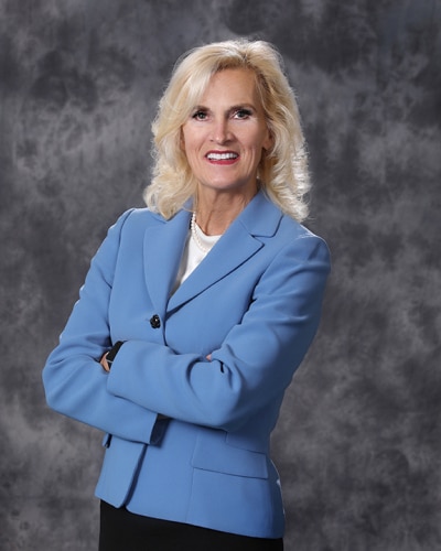 A woman with shoulder-length blonde hair, wearing a light blue blazer and white top, stands with arms crossed and smiles at the camera against a gray mottled background.