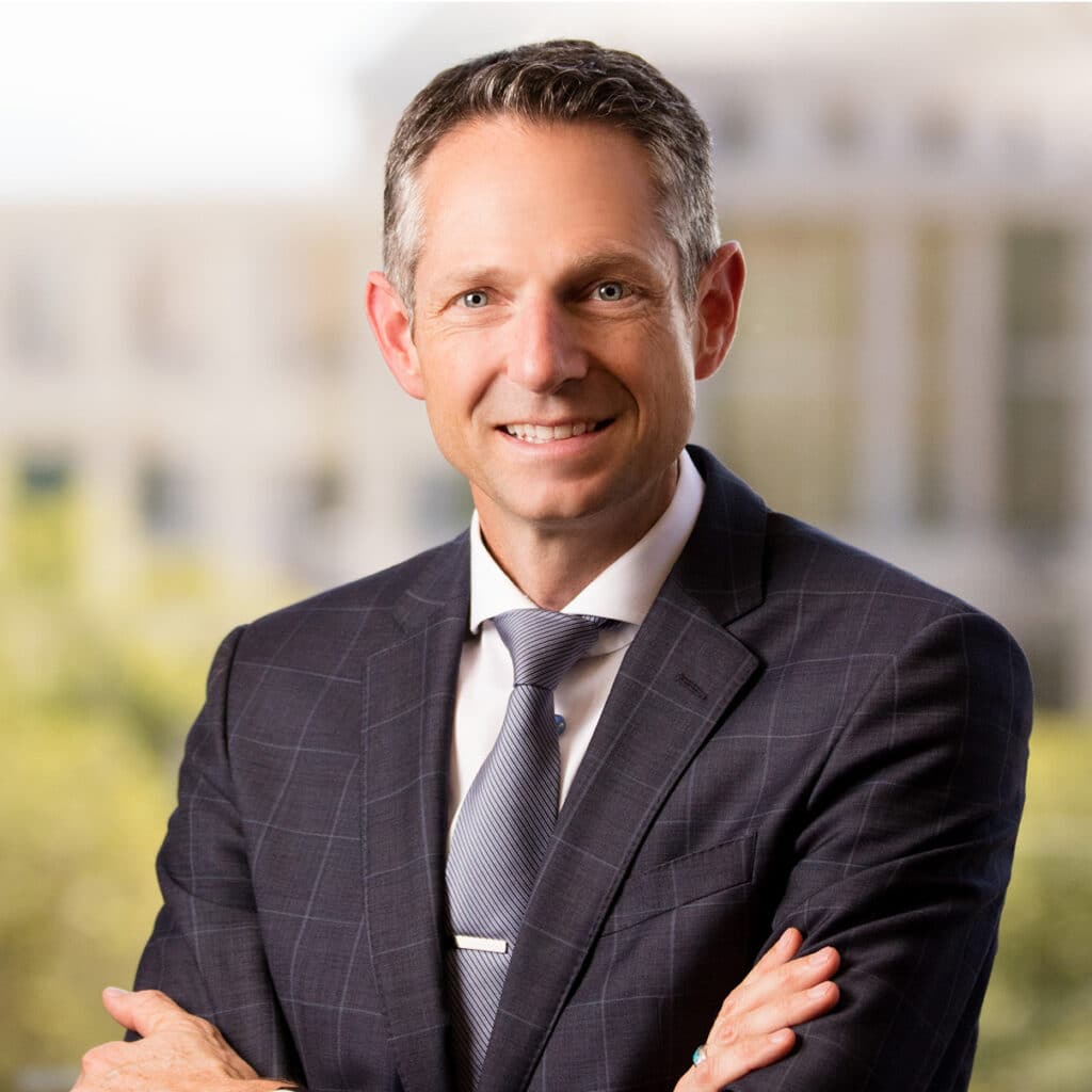 A man in a dark checked suit and tie stands with arms crossed, smiling confidently in front of a blurred office building background.