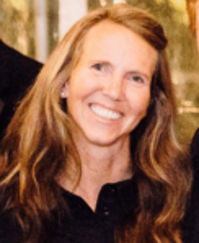 A woman with long, wavy light brown hair and fair skin smiles at the camera. She is wearing a black top and is standing indoors with a blurred background.