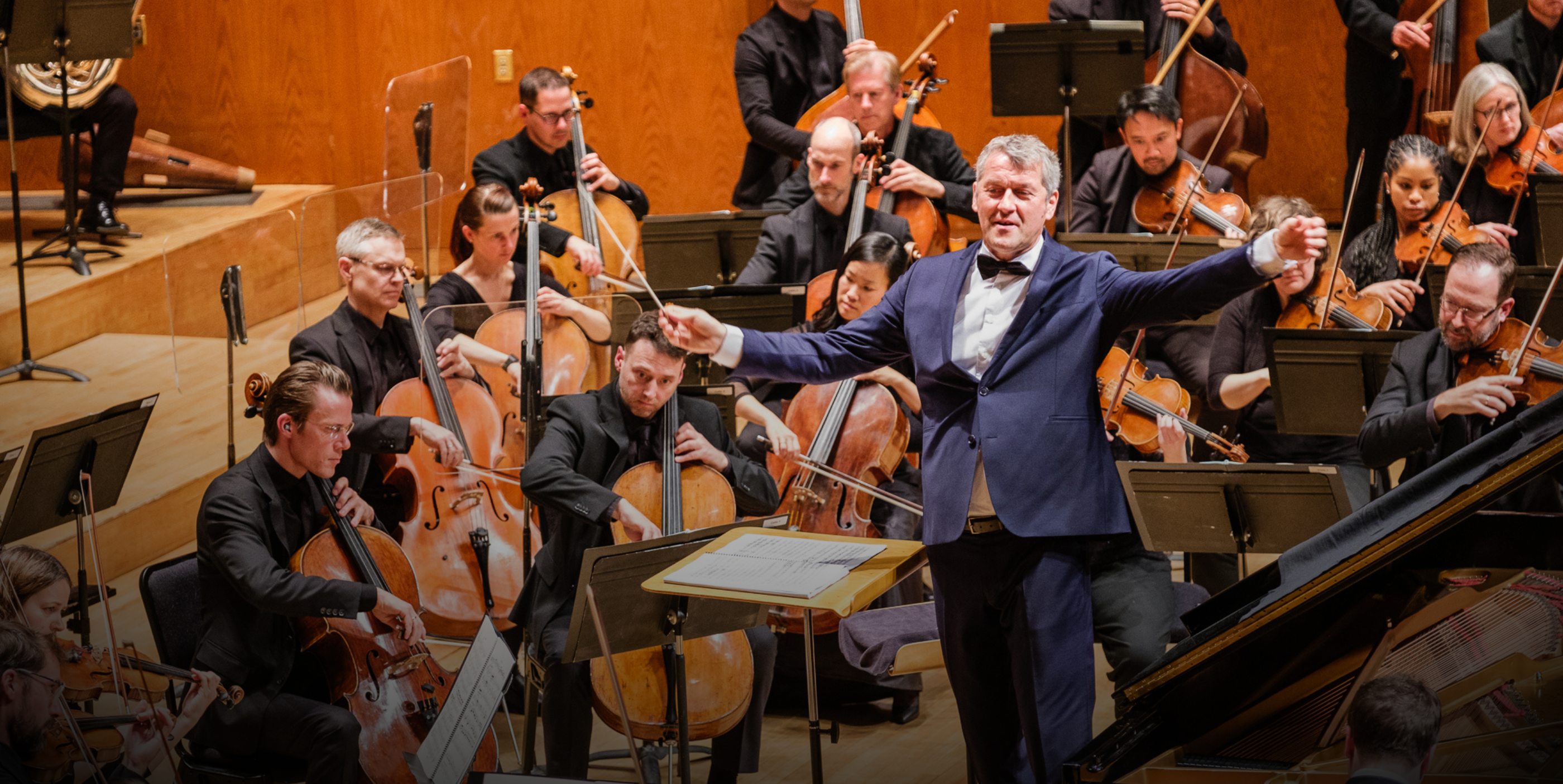 A conductor in a blue suit leads an orchestra of musicians playing string instruments on a wooden stage, with music stands and sheet music visible.