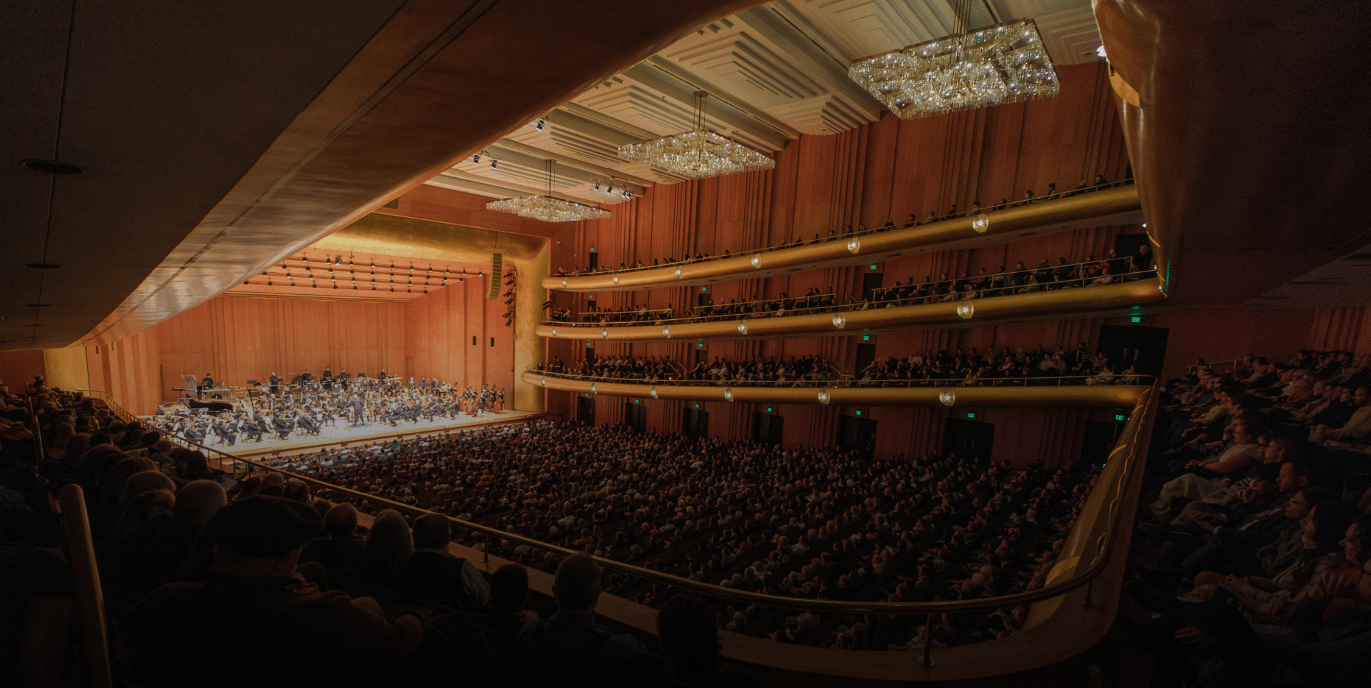 A large, elegant concert hall filled with an audience watching an orchestra perform on a well-lit stage, with balconies and chandeliers accenting the grand interior.