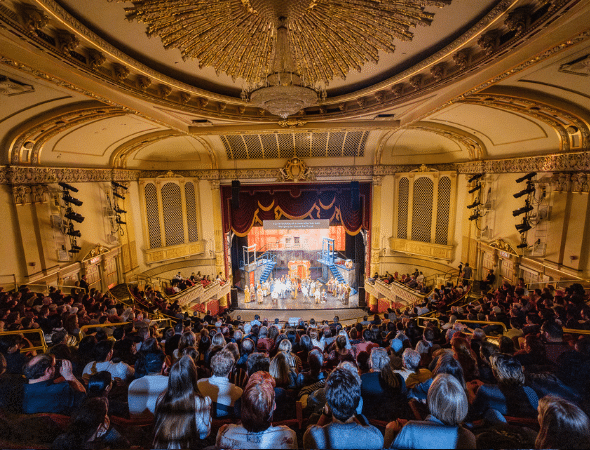 A large audience sits in an ornate theater with a crystal chandelier, watching a colorful stage performance. The theater features gold accents, arched ceilings, and balconies filled with people.